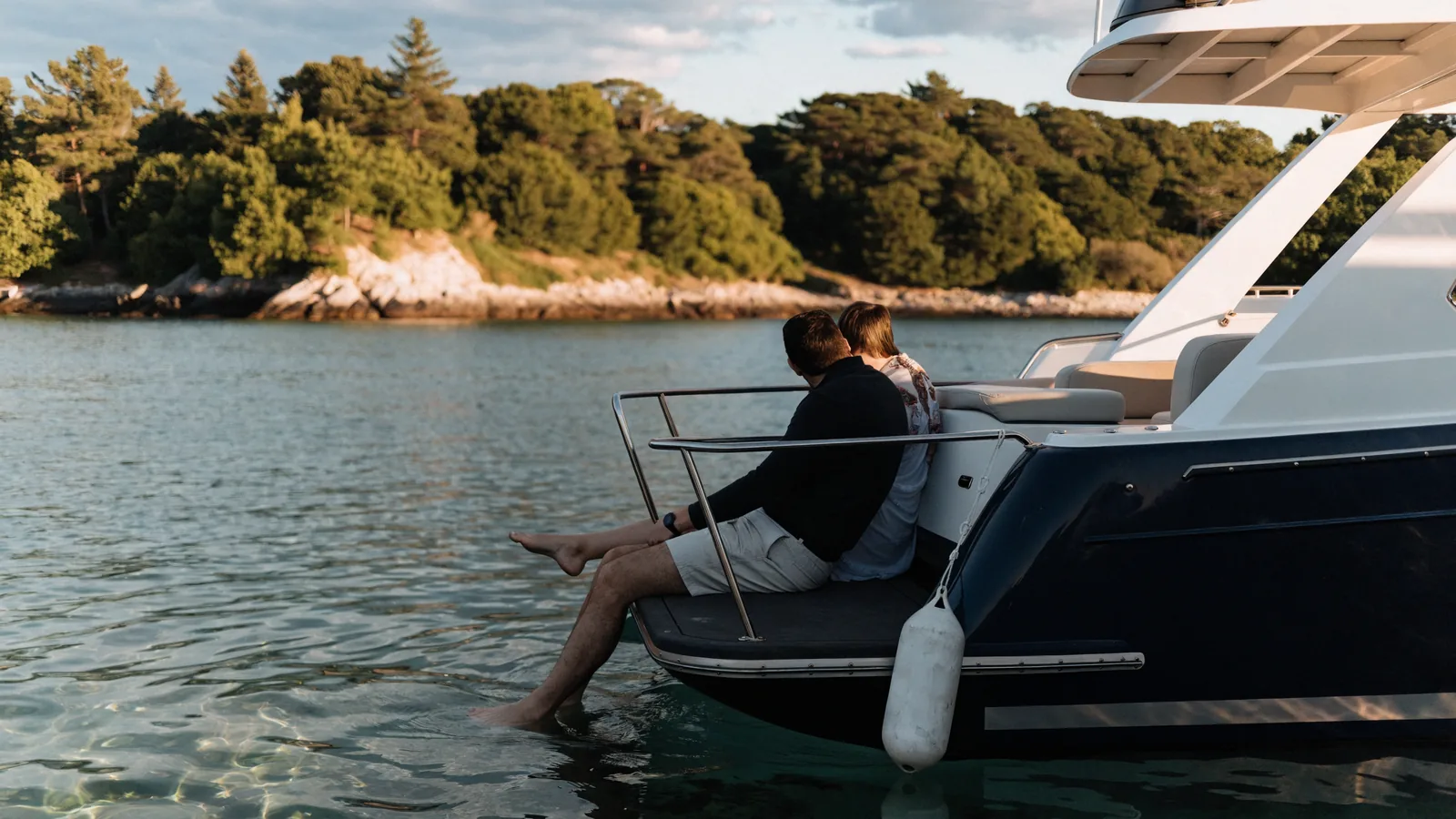 Ein Paar sitzt am Abend auf der Badeplattform seiner Motoryacht in einer ruhigen Bucht, die Füße im Wasser