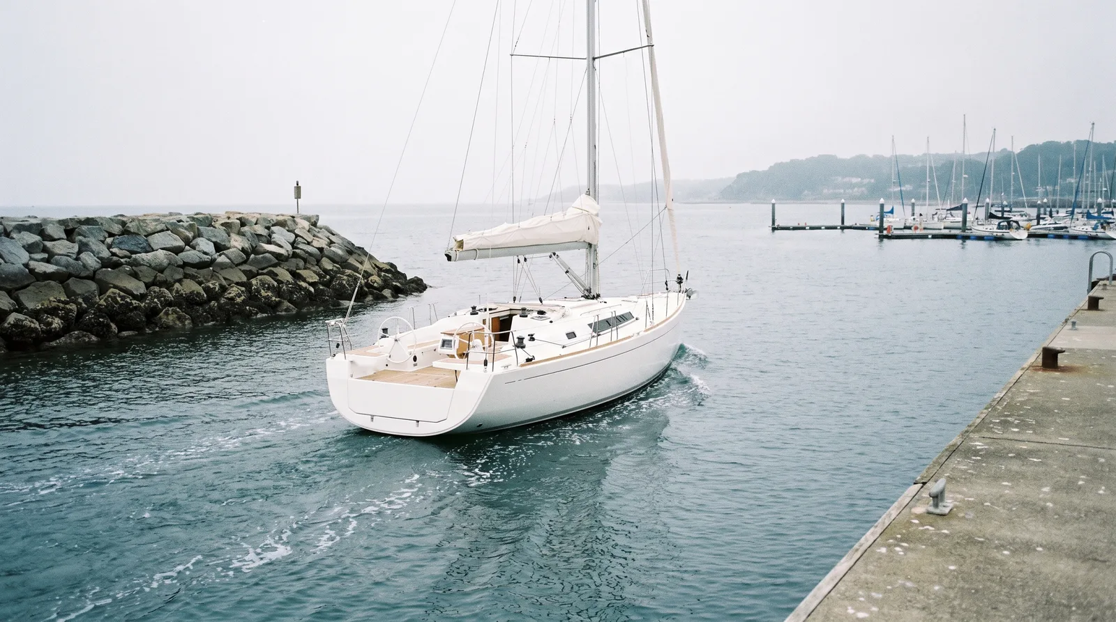Weiße Segelyacht mit geborgenen Segeln fährt unter Maschine aus dem Hafenbecken, links die Mole aus Felsblöcken, rechts eine Betonkaje, hinten Stege im Dunst.