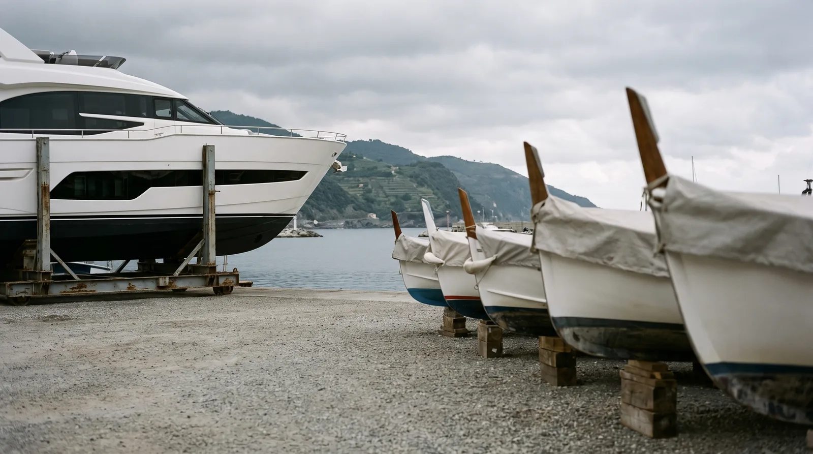Werftgelände an der Küste Liguriens: links eine neue weiße Motoryacht auf einem Stahlgestell für den Transport, rechts eine Reihe kleiner Boote mit hellgrauen Planen auf Holzklötzen, im Hintergrund das ruhige Hafenbecken und grüne Terrassenhänge.