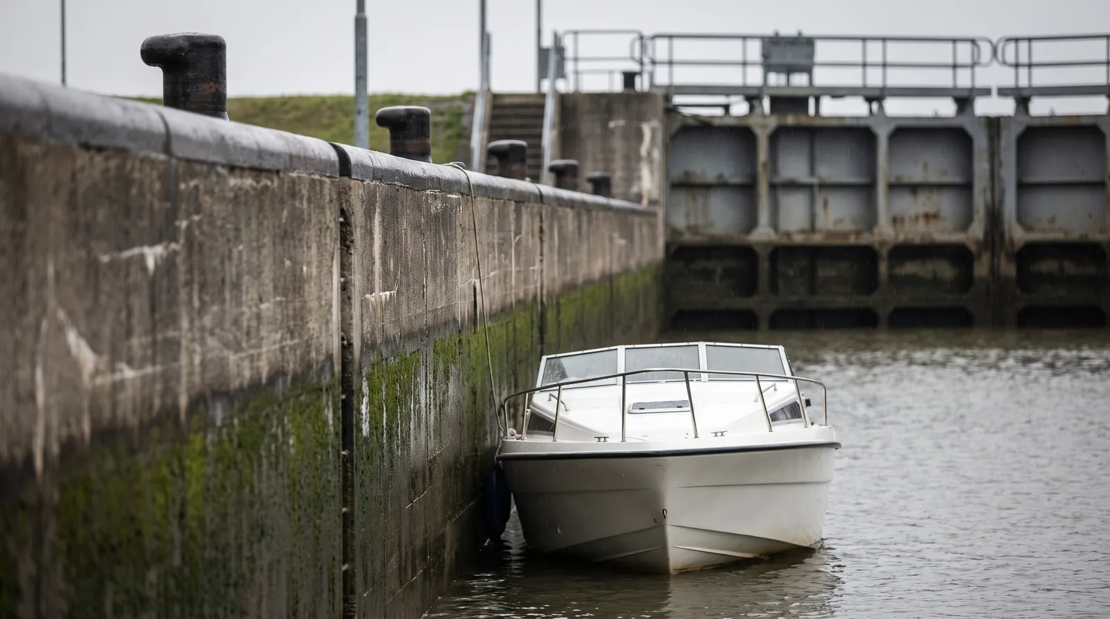 Ein weißes Motorboot wartet an der nassen Betonwand einer Schleusenkammer, an der Kante stehen eiserne Poller, dahinter liegt das Schleusentor im Dunst.
