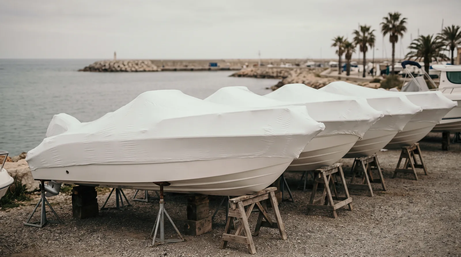 Neue, in weiße Folie eingeschweißte Motorboote stehen nebeneinander auf Holzböcken auf einem Schotterplatz am Wasser, im Hintergrund Wellenbrecher und Palmen.