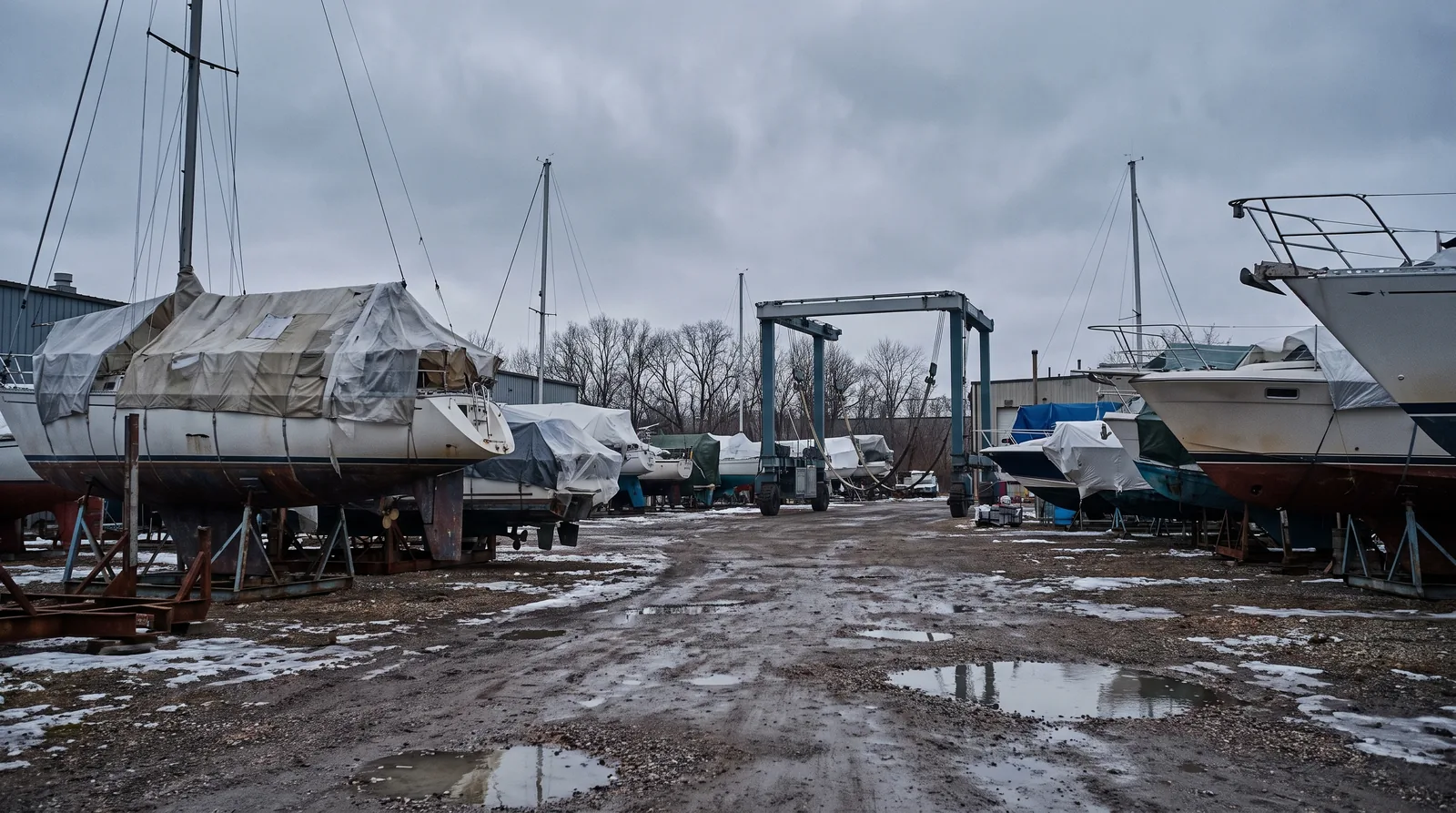 Boote stehen im Winterlager an Land auf Böcken unter Planen, im Hintergrund ein Travellift