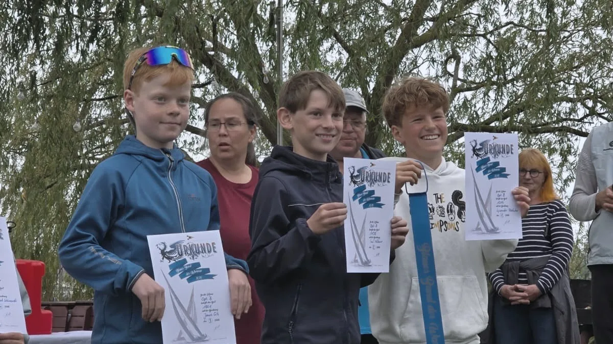Drei Jugendliche mit Segel-Urkunden am Steg, einer mit dem blauen Band der Regatta um den Hals, Trauerweide im Hintergrund