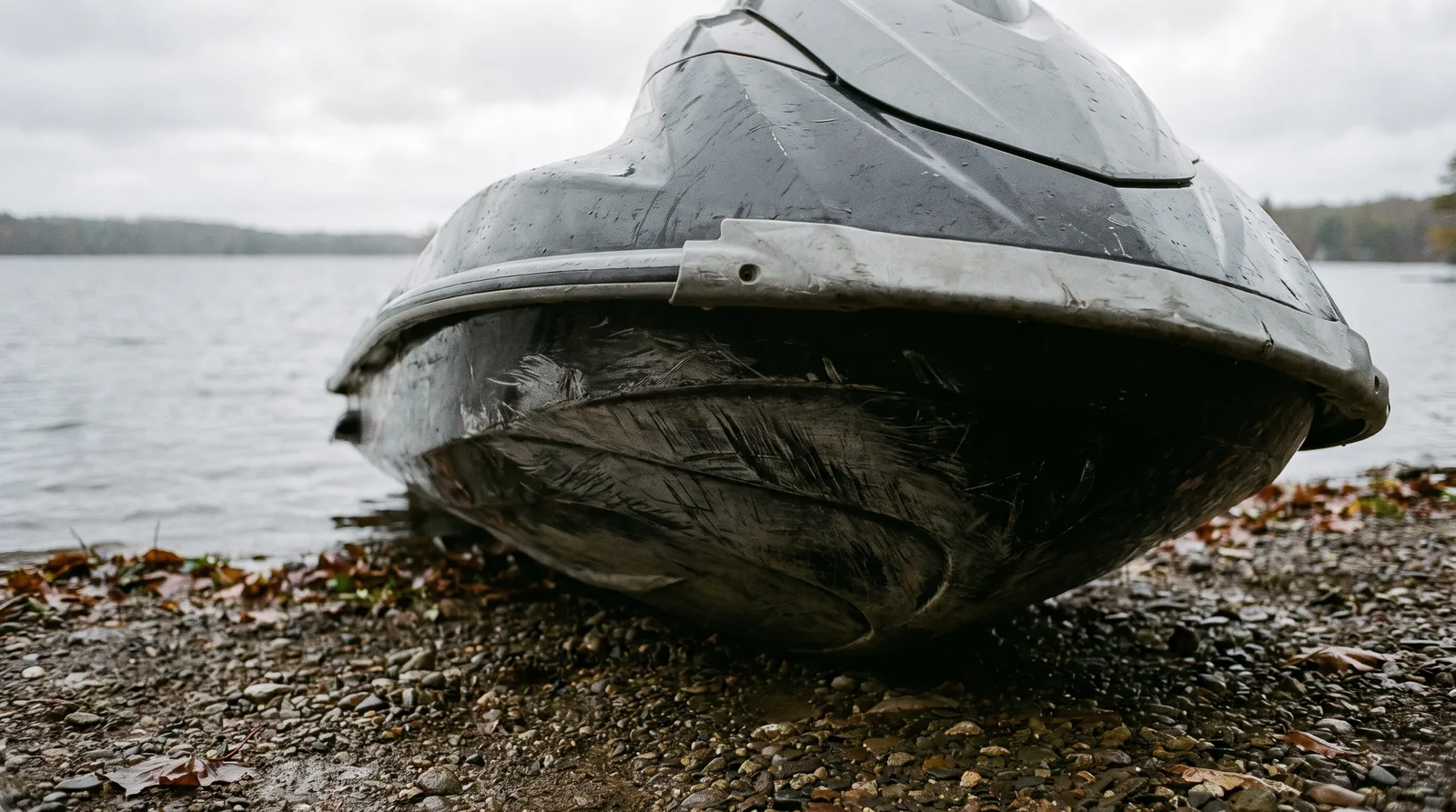Bug eines Jetskis mit Kratzern am Rumpf, an einem Kiesufer liegend, bedeckter Himmel.