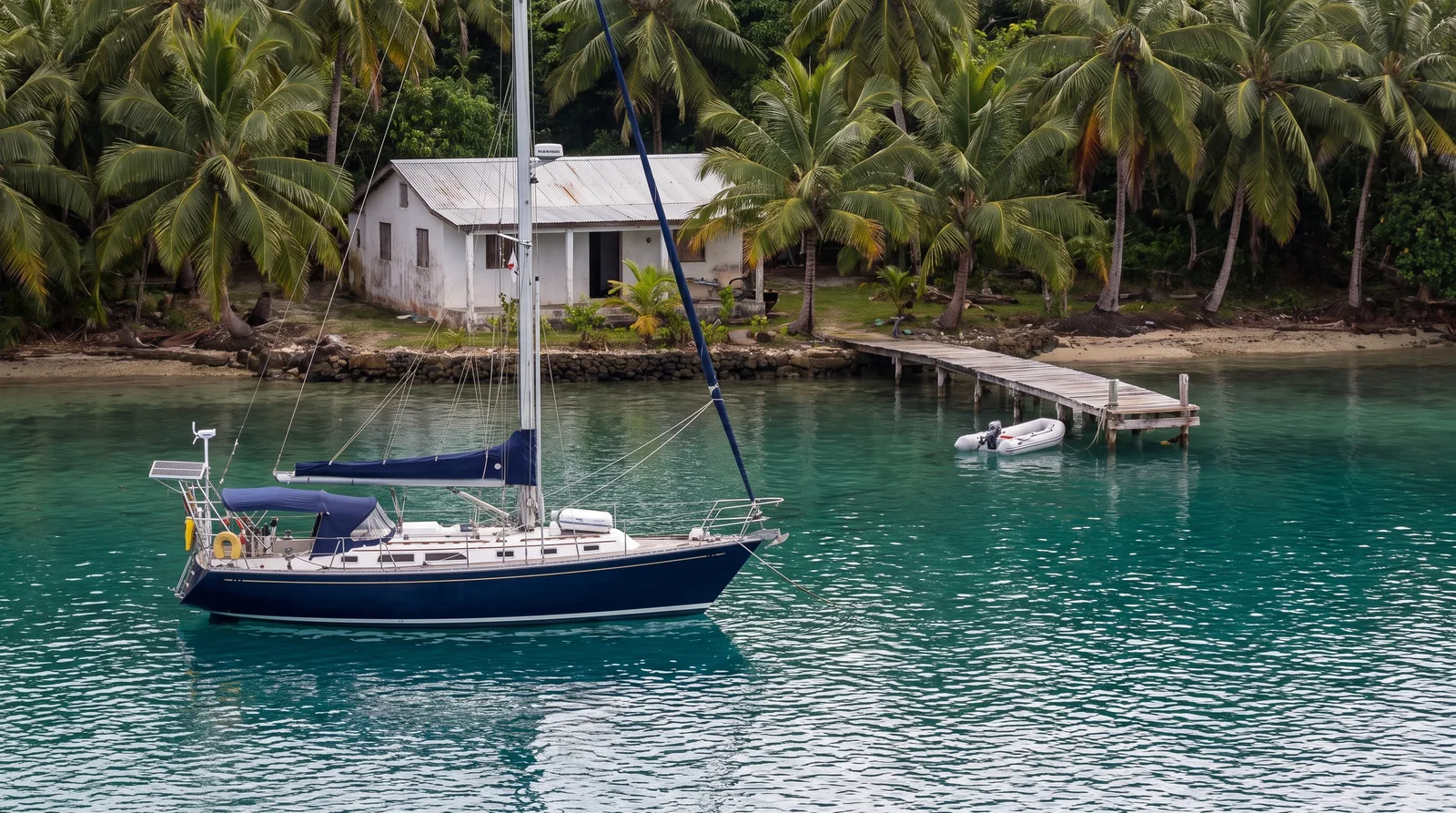 Fahrtenyacht ankert in einer tropischen Bucht, am Ufer steht eine kleine Klinik
