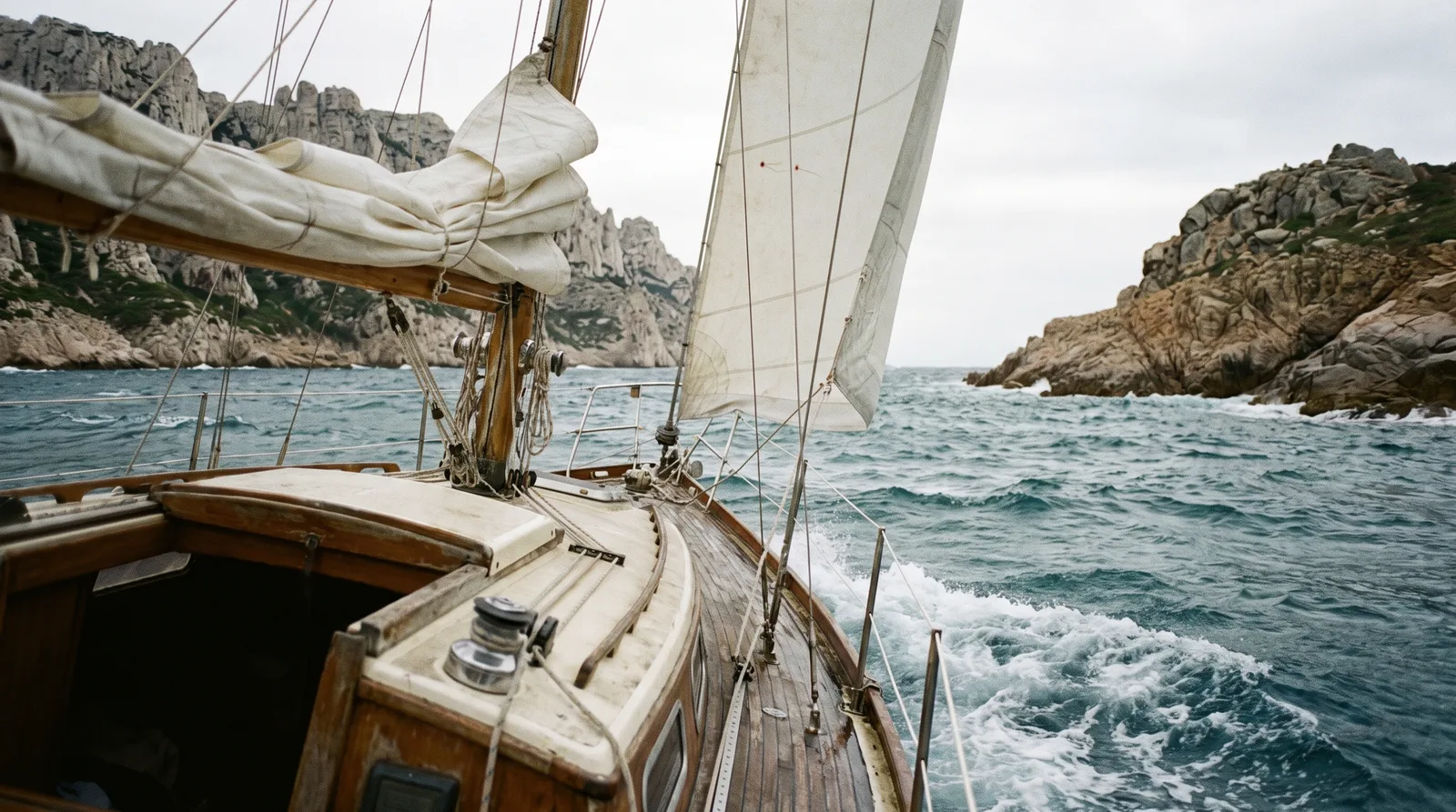 Gepflegte Holzsegelyacht mit gerefftem Vorsegel quert bei kräftigem Wind und Schaumkronen eine Meerenge zwischen Kalkklippen und Granitfelsen