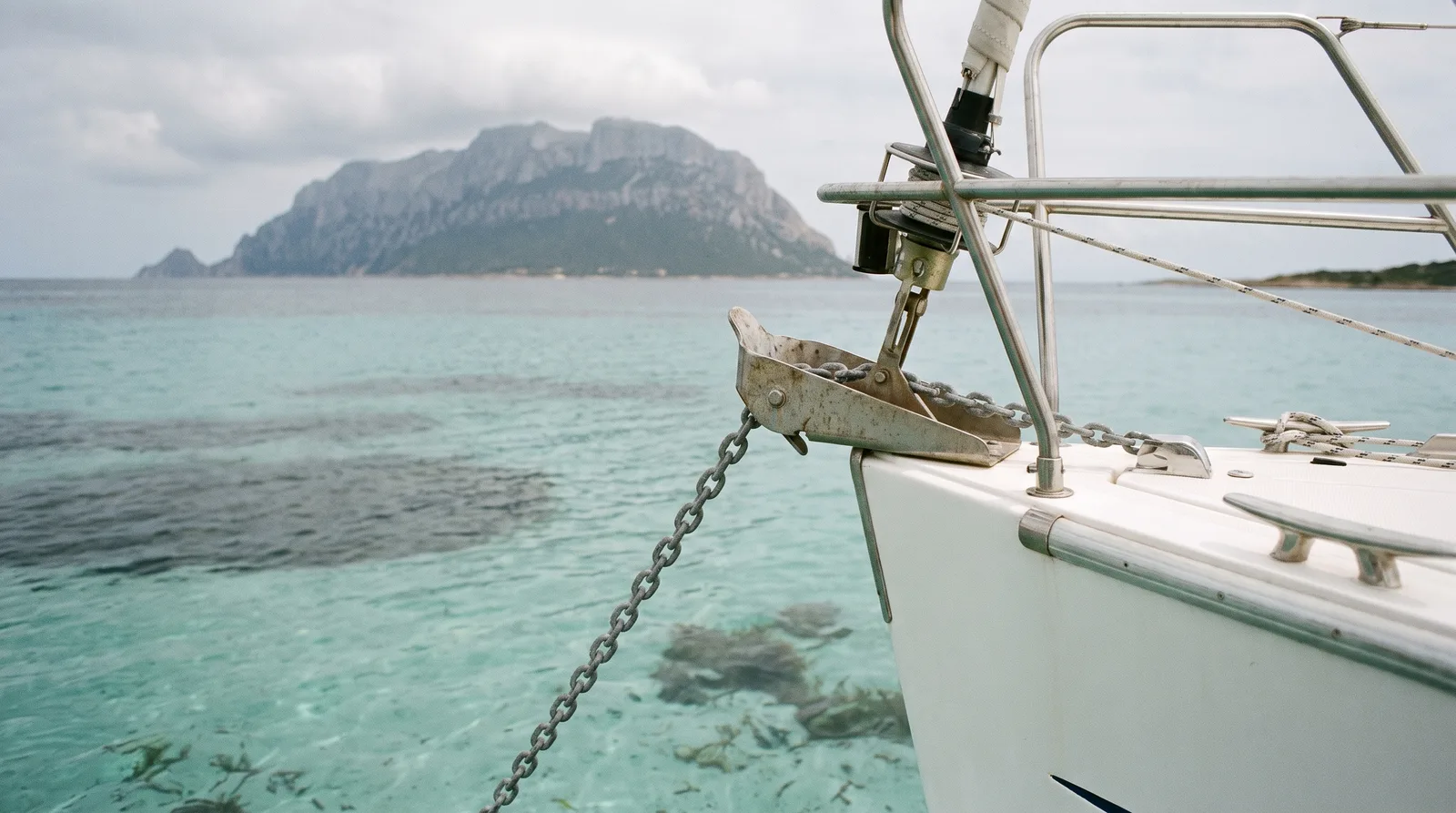 Bug einer Segelyacht mit Ankerkette über hellem Sand, im Hintergrund die Insel Tavolara