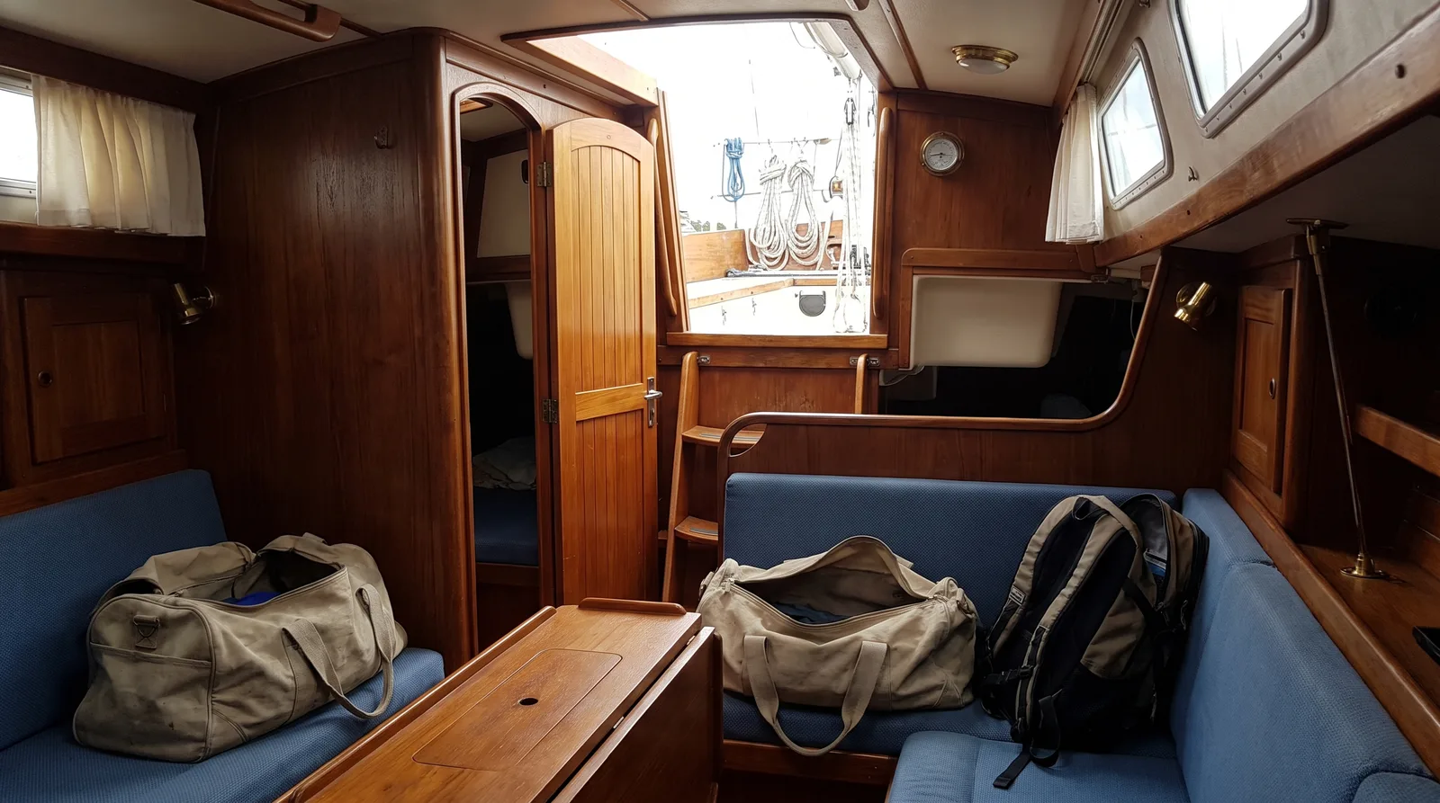 Salon einer Segelyacht mit Holzausbau, auf den Polsterbänken offene Reisetaschen und ein Rucksack, der Niedergang offen