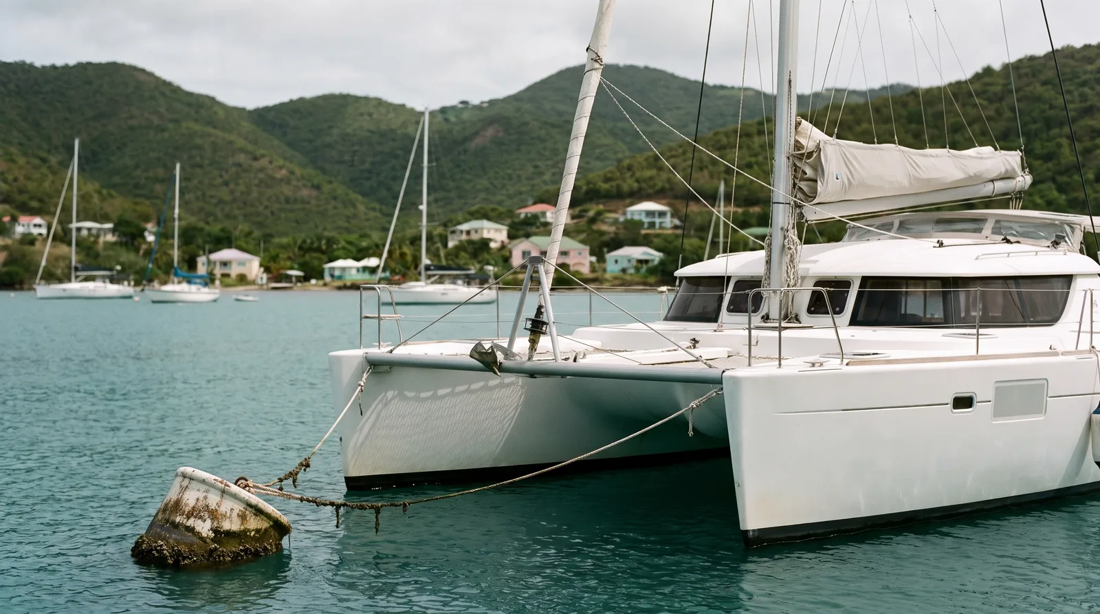 Weißer Segelkatamaran an einer Boje in einer ruhigen karibischen Bucht, dahinter grüne Hügel mit kleinen bunten Häusern und weitere Yachten