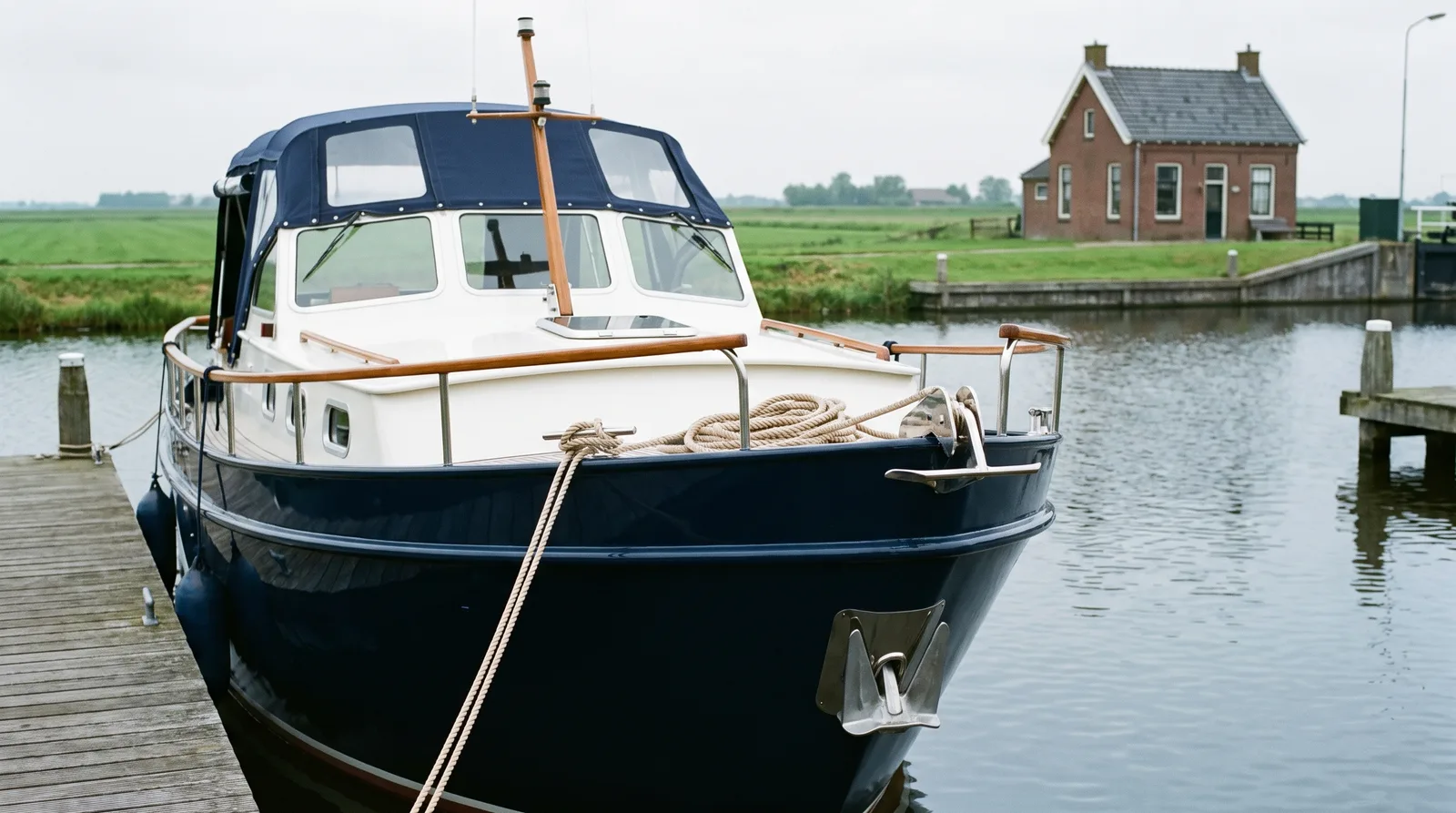Gepflegter Stahlkreuzer mit blauem Verdeck an einem friesischen Hafen am IJsselmeer, dahinter flaches Polderland und ein Schleusenhaus aus Backstein
