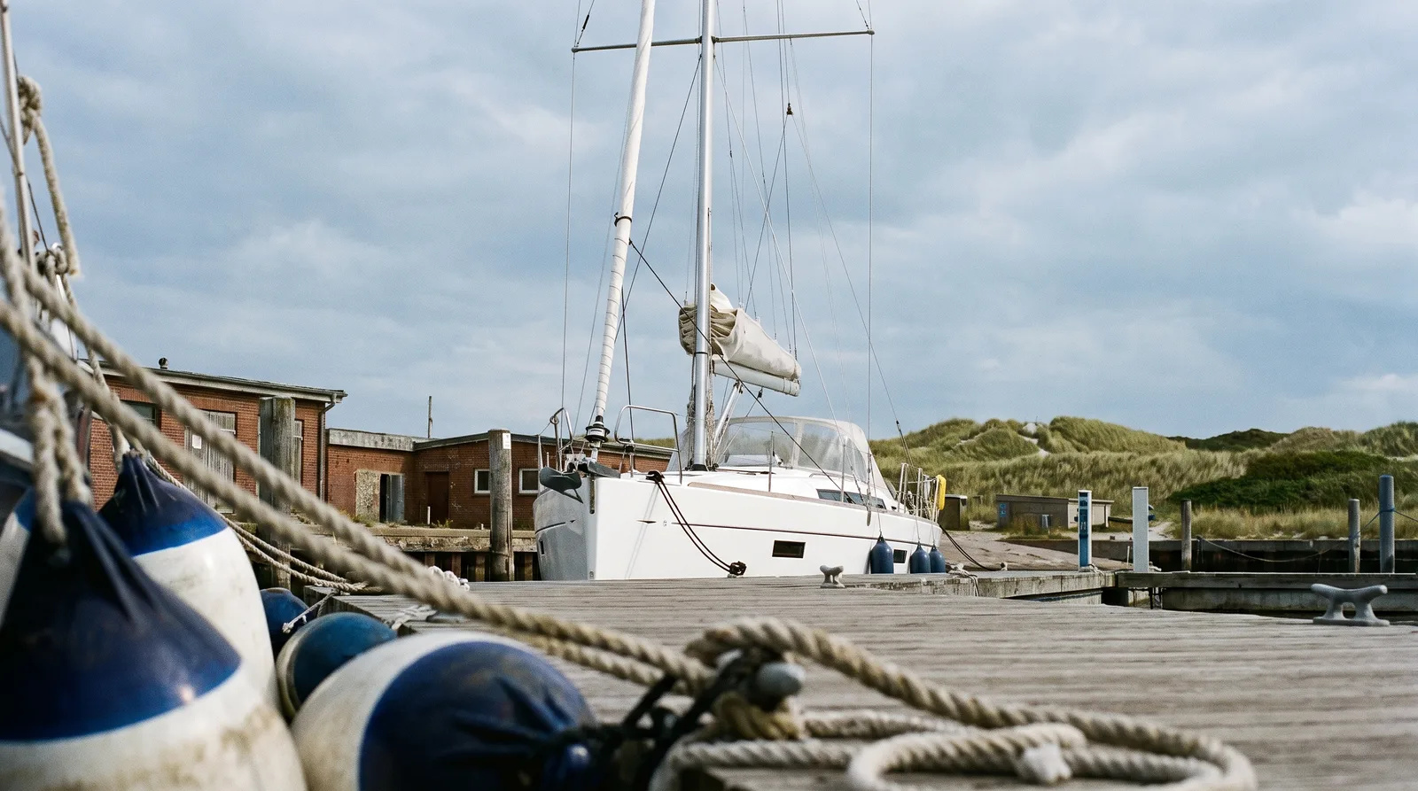 Segelyacht an einem Holzsteg in einem Ostseehafen, vorn Fender und Leinen, dahinter Backsteinhäuser und Dünen