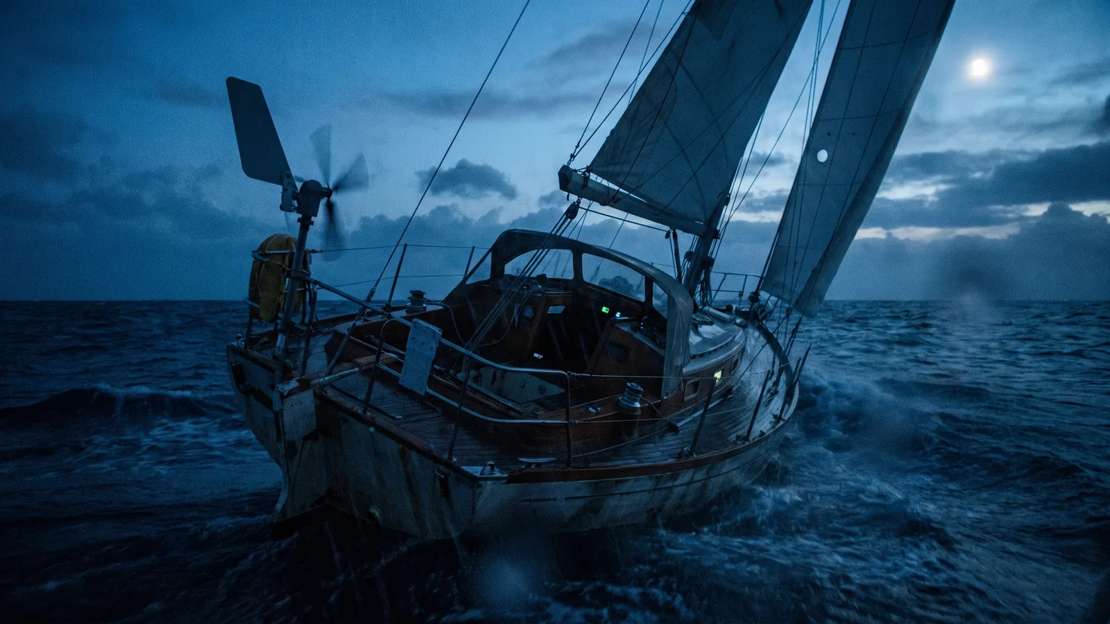 Eine Segelyacht fährt nachts unter Windsteueranlage, das Cockpit ist leer, am Horizont die Lichter der Adriaküste