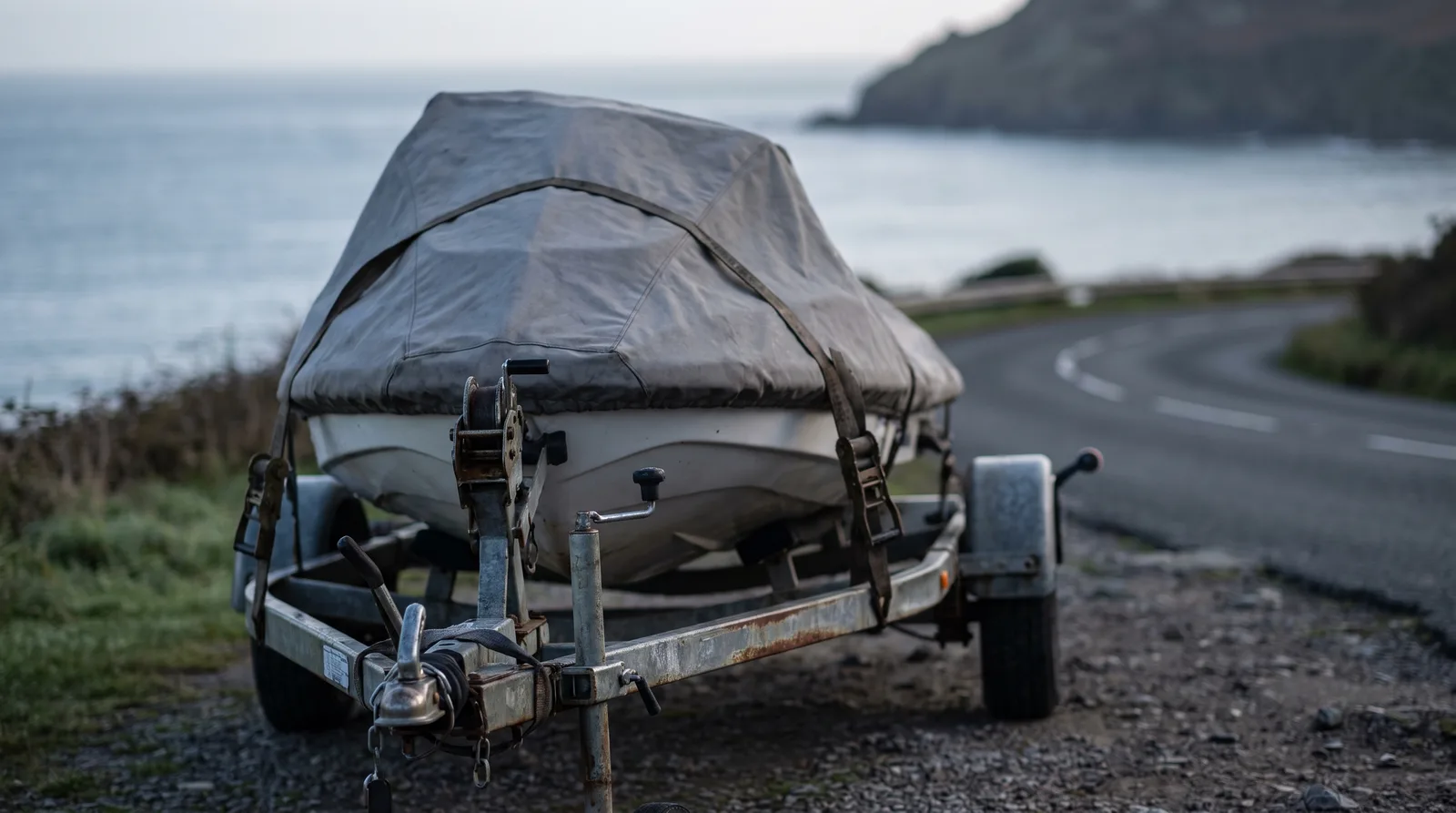 Ein abgedecktes Wassermotorrad festgezurrt auf einem Trailer an einer Küstenstraße, im Hintergrund das Meer