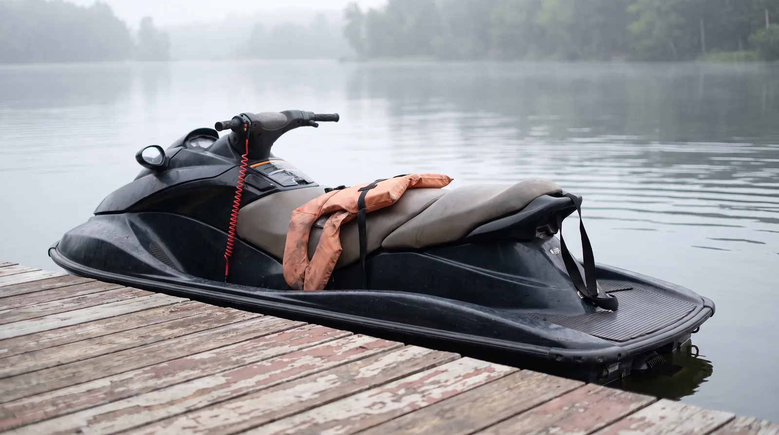 Ein leerer Jetski mit Schwimmweste auf der Sitzbank und roter Notstoppleine am Lenker an einem Holzsteg im Morgendunst