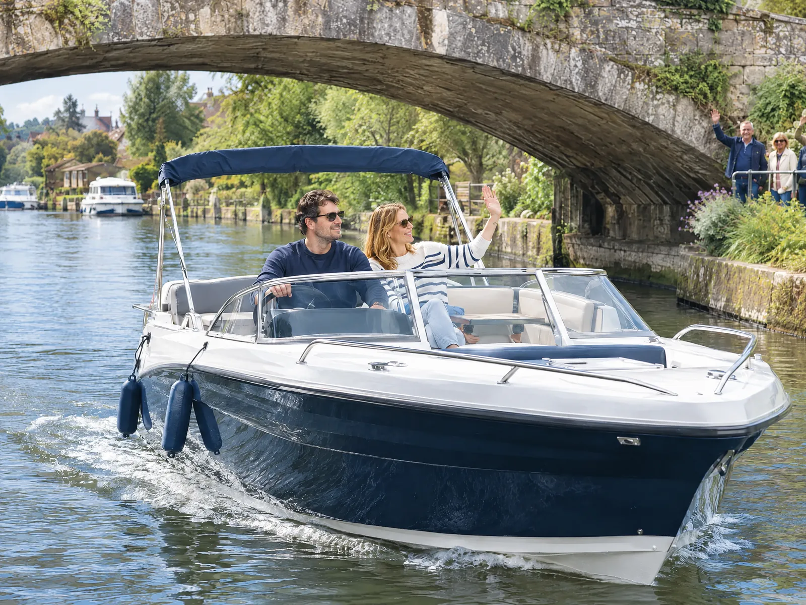Ein Paar fährt mit dem Motorboot unter einer alten Steinbrücke durch, sie winkt den Leuten am Ufer zu