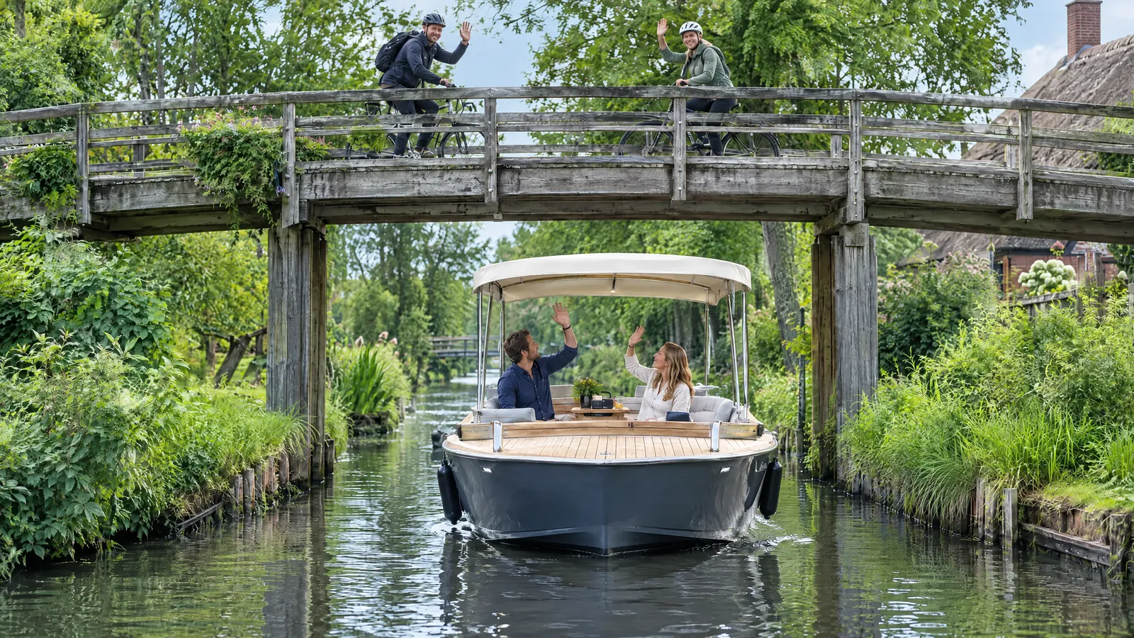 Ein Paar winkt aus einem Elektroboot mit Sonnendach zwei Radlern auf einer Holzbrücke zu