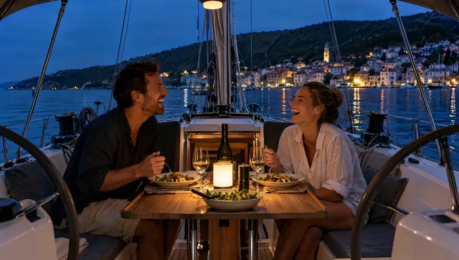 Ein Paar lacht beim Abendessen im Cockpit einer Segelyacht, am Ufer spiegeln sich die Lichter eines Städtchens